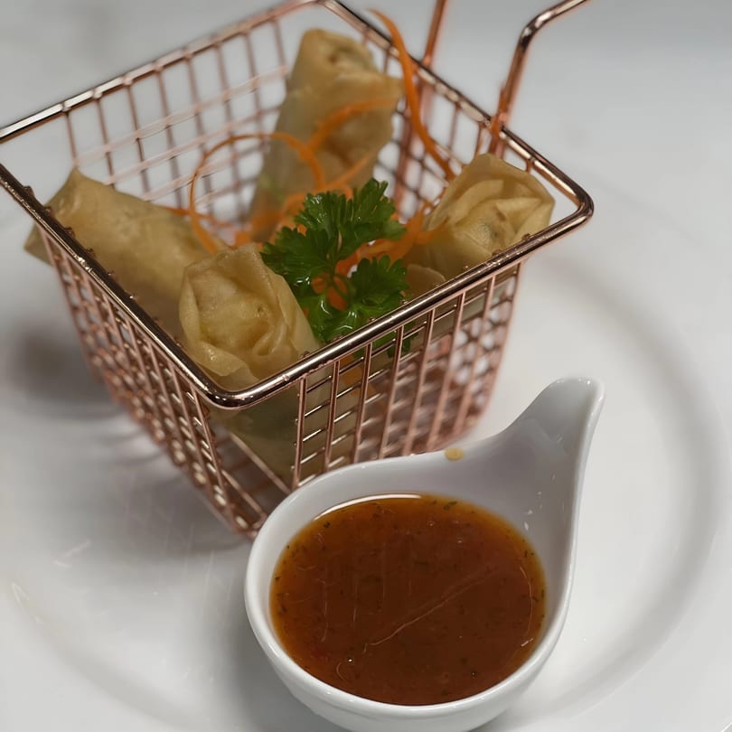 Crispy spring rolls in a rose gold basket with parsley, carrot strips, and a side of dipping sauce.