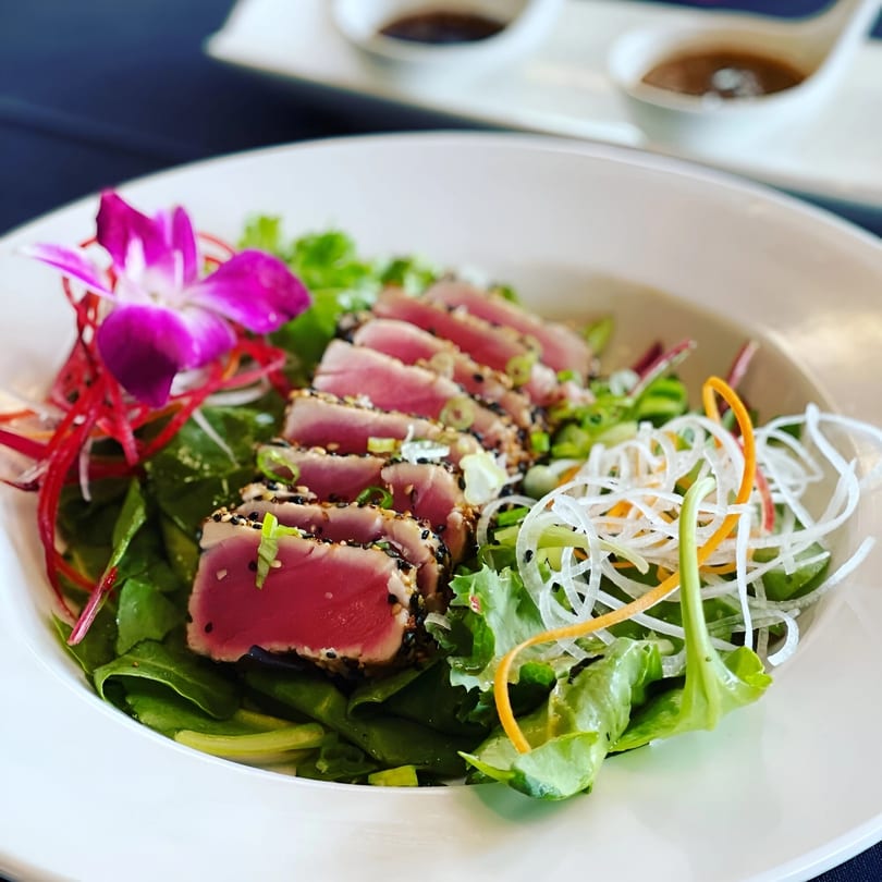 Seared tuna salad with greens, shredded vegetables, and a purple orchid garnish in a white bowl.